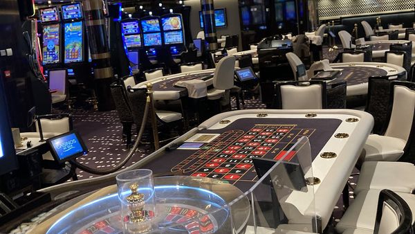 Empty shipboard casino with a roulette table and rows of lit slot machines on a cruise ship