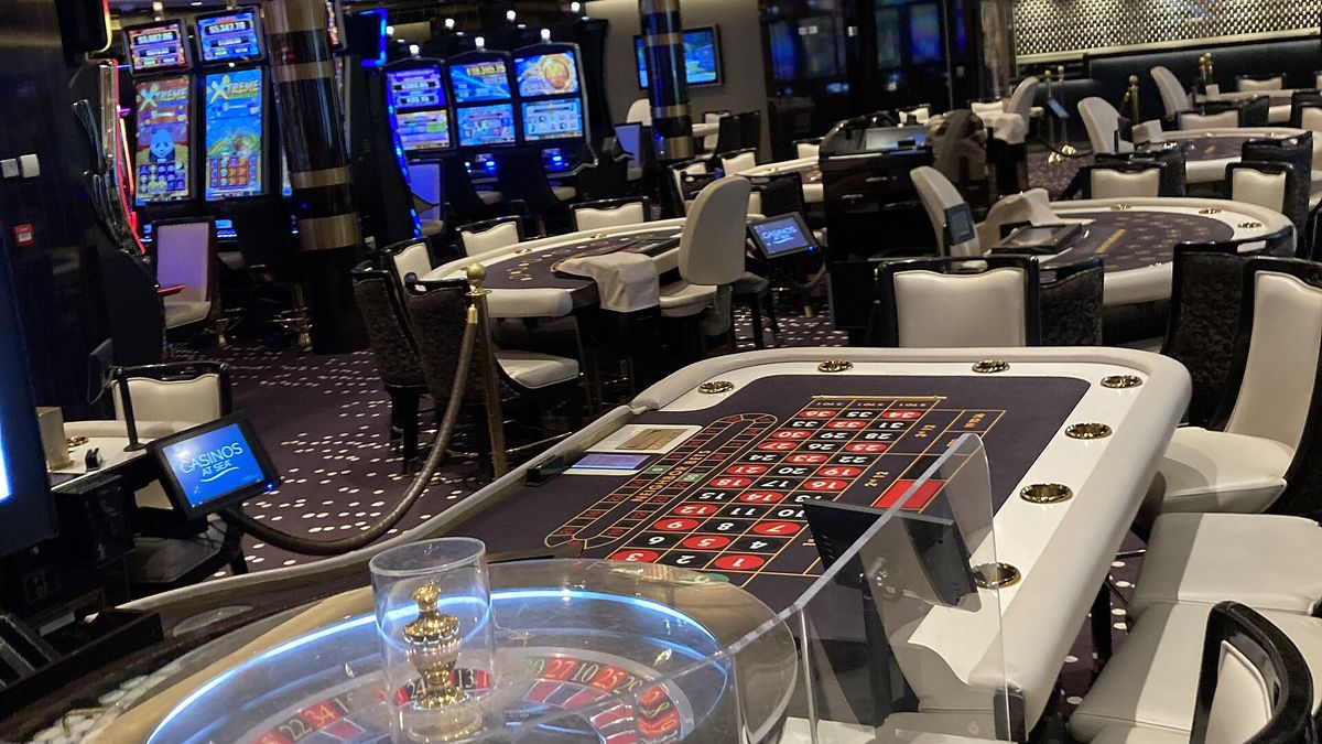 Empty shipboard casino with a roulette table and rows of lit slot machines on a cruise ship